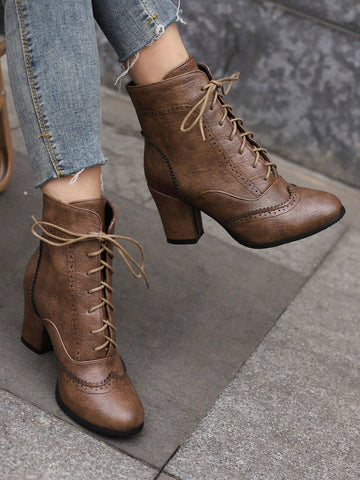 A pair of brown lace-up ankle boots with block heels and brogue detailing, shown worn with frayed blue jeans on a gray stone pavement.