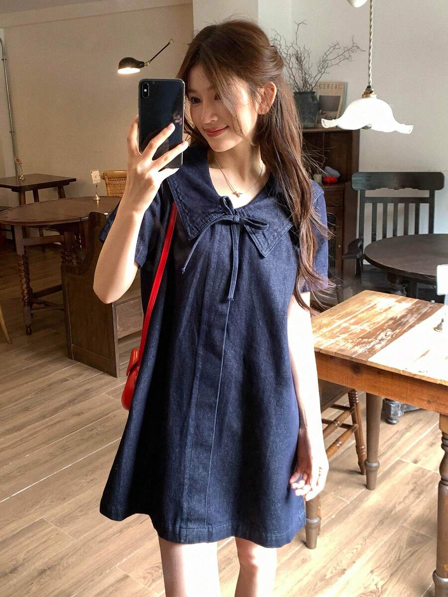 Three-quarter front view of dark-indigo sailor-collar denim mini dress with chest bow and short sleeves