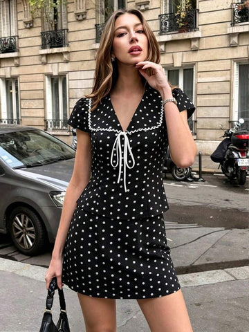 Front city view of woman in black polka dot blouse and matching high-waisted mini skirt set with white collar trim and tie