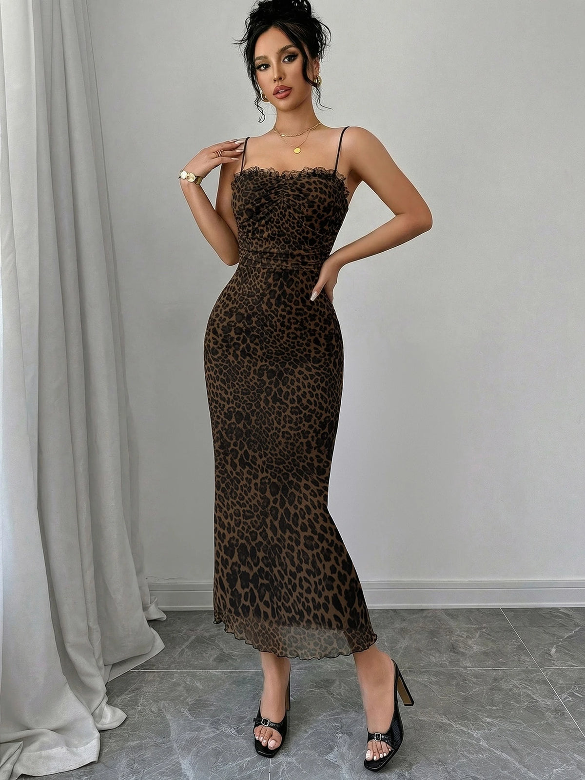 A woman models a brown and black leopard print spaghetti-strap maxi dress with a lace-trimmed sweetheart neckline, styled with black strappy heels against a gray wall and draped white fabric.
